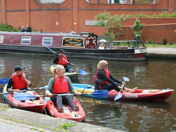 A landscape image showing 4 people sat in kayaks laughing. Behind them is a heritage working canal boat.