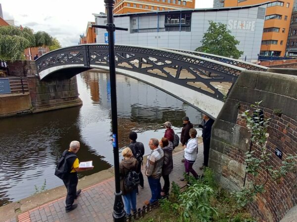 A landscape showing a group of people stood on a canal towpath under an arched bridge. They are being led by someone in a yellow shirt.