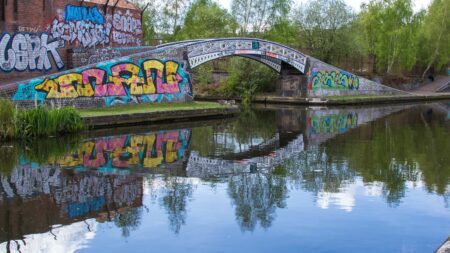 A landscape showing an arched bridge covered in colourful graffiti. Behind that a are green trees. All the bridge, the graffiti and bridge is reflected in the canal water below.