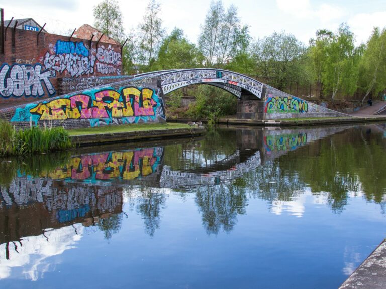 A landscape showing an arched bridge covered in colourful graffiti. Behind that a are green trees. All the bridge, the graffiti and bridge is reflected in the canal water below.