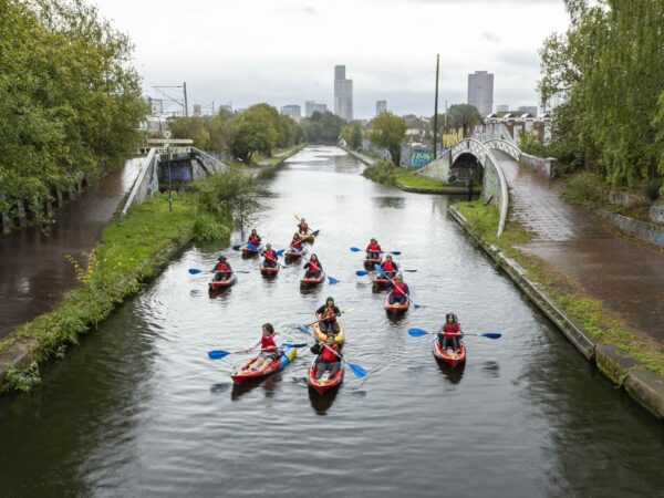A landscape showing a group of kayakers on a canal. Either side of them are arched bridges and paths. Behind them are large skyscrapers.