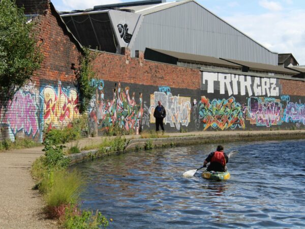 A landscape showing a kayaker on curve of the canal. There is a person walking on the path next to the canal and behind them is a lot of graffiti.