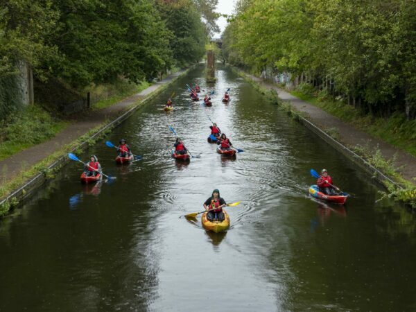 A landscape image showing a group of kayakers paddling towards the camera. The canal is lined with towpaths and lots of green trees. The kayakers are being led by someone in a yellow and orange kayak.