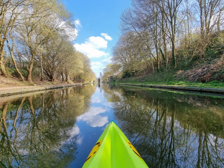 A landscape image showing the front of a neon green kayak. The canal is lined with towpaths and green grass, trees and a blue sky above, all of which is reflected in the canal water.