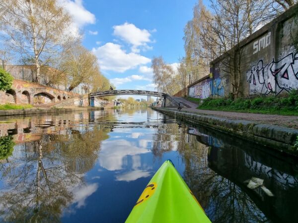 A landscape image showing the front of a neon green kayak. To the right is a wall with graffiti and in the distance is an arched bridge. The sky and landscape is reflected in the canal water.