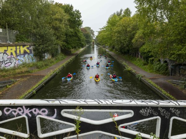 A landscape image showing a group of kayakers on a canal lined with green trees and weeds. In the foreground is a black and white metal bridge with graffiti on and to the left is a wall with graffiti on.
