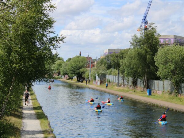 A landscape image showing some kayakers on a canal. To the left is a path with people walking on. In the distance are the tops of buildings and a crane.