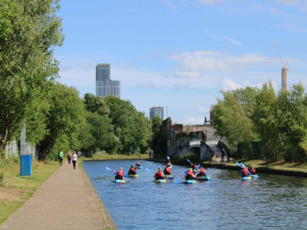 A landscape image showing a group of 11 kayakers on a canal. To the left of the image is a path with people walking down.