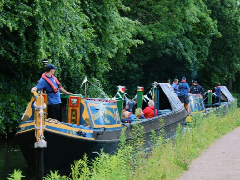 A landscape image showing a two canal boats on a canal. Behind them are a lot of green foliage.