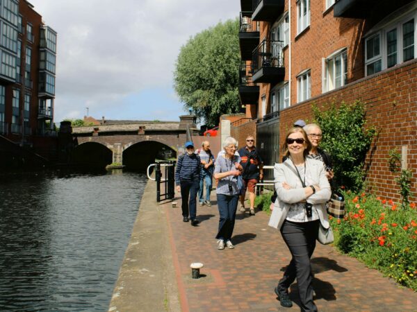 A landscape image showing a group of people walking along a canal towpath towards the camera. The canal is to the left of the image.