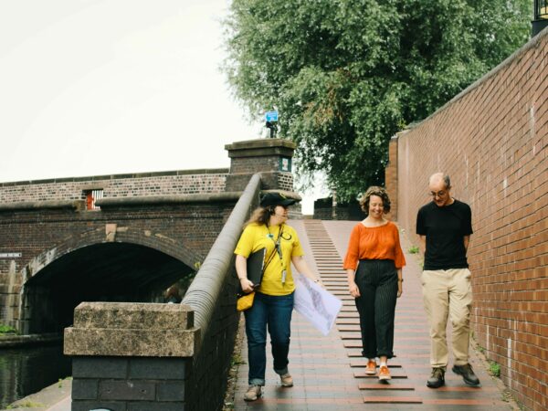 A landscape image showing three people walking down a ramp onto a canal towpath. There is an arched bridge to their left.