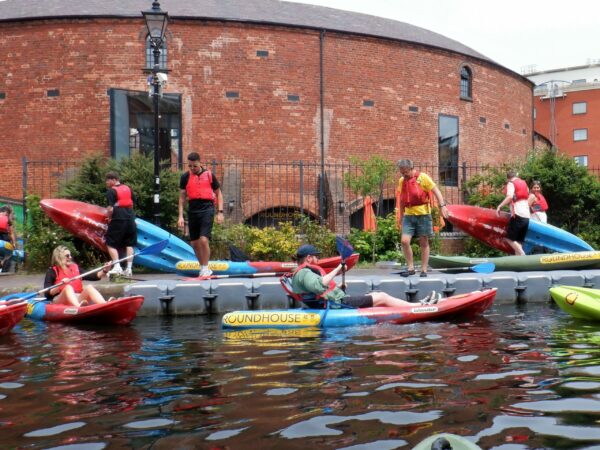 A landscape image showing a group of people getting into kayaks on the canal water. Some people are stood on a towpath behind. Behind everyone is a red bricked curved building.