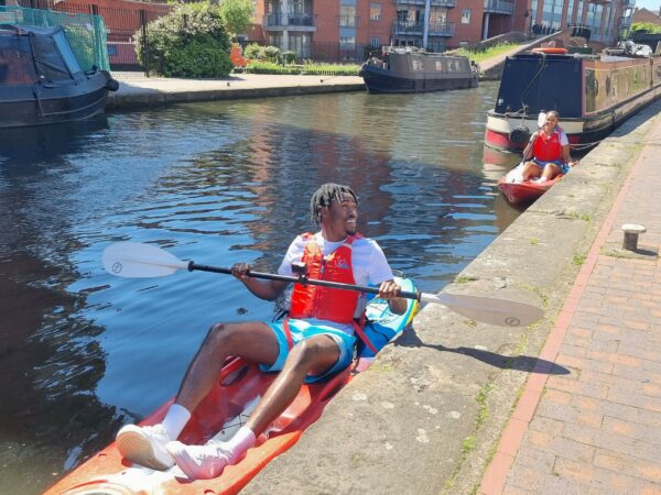 A landscape image showing two people sat in kayaks on the canal, one in front of the other. They are next to the path and holding oars, with canal boats around them.