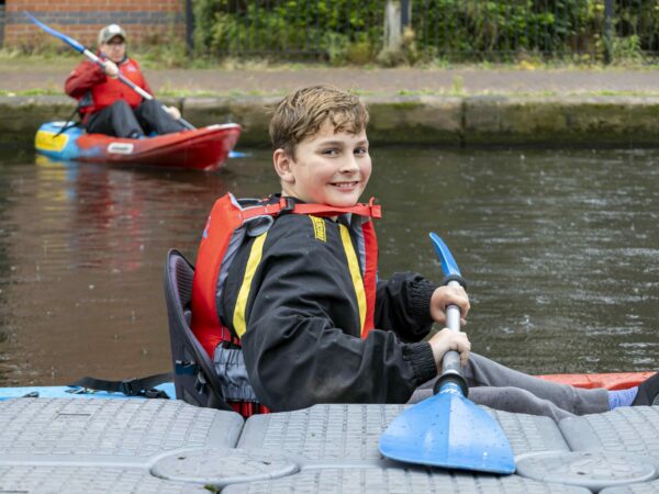 A landscape image showing a young person holding a blue ended oar, smiling at the camera. Behind them is a person paddling on a kayak on the canal water.