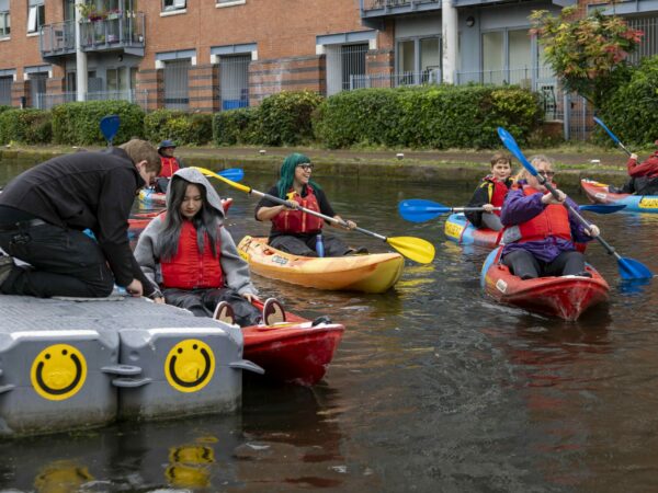 A landscape image showing multiple people in kayaks on the canal water. Someone dressed all in black is knelt down on a grey pontoon helping someone into a kayak.