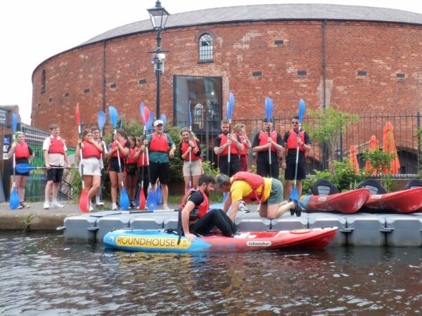 A landscape image showing a person in a yellow t-shirt helping someone into a blue and red kayak on the canal. Stood watching on a towpath are a large group of people all holding oars upright. Behind them is a red bricked curved building.