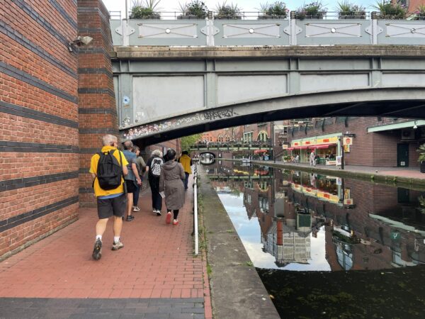 A landscape image showing a group of people walking on a red tiled path under an arched bridge. To their right is a canal, the waters reflecting all the shops on the wharf in it.