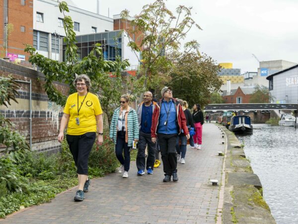 A landscape image showing a group of people walking along a towpath with the canal to the right of the image. They are being led by someone in a yellow t-shirt.