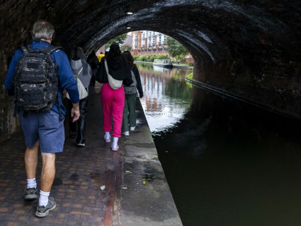 A landscape image showing a group of people walking along a towpath under an arched bridge, with the canal to their right.