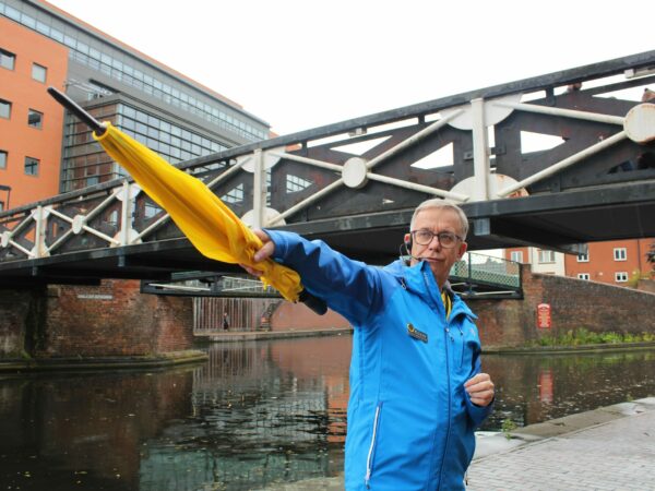 A landscape image showing a person in blue pointing a yellow umbrella. Behind them is a black and white metal bridge and a canal.