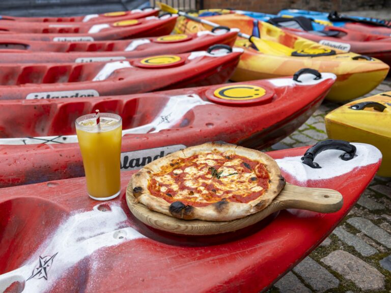 A landscape image showing a row of red kayaks with a cheese and tomato pizza on a wooden board on it. There is also a pint of orange juice with a red and white striped straw.