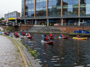 A landscape image showing a group of kayakers on a canal. Behind them is a set of stairs and a large windowed building.