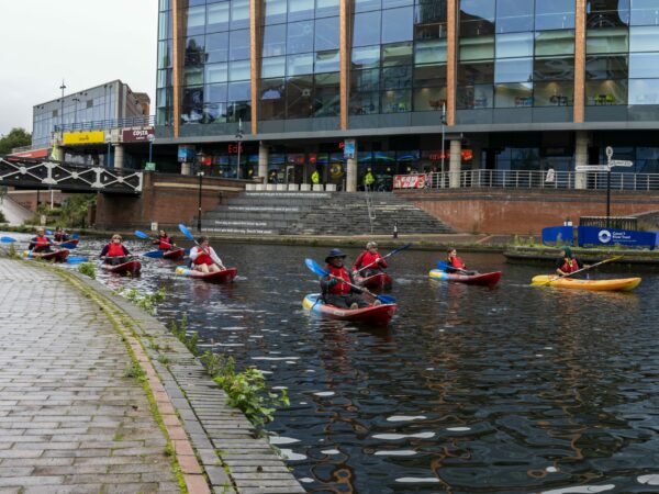 A landscape image showing a group of kayakers on a canal. Behind them is a set of stairs and a large windowed building.
