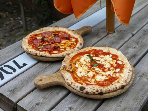 A landscape image showing two pizzas on wooden paddles on a wooden table.