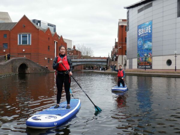 A landscape image showing two people doing stand up paddle boarding on canal water.