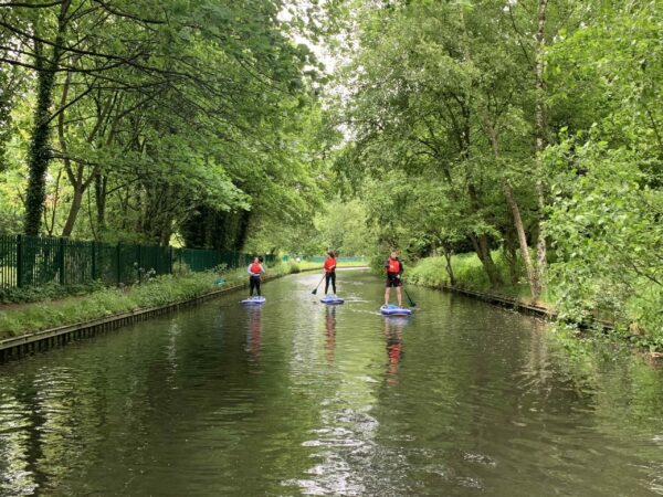 A landscape image showing three people doing stand up paddle boarding on a canal which is lined with green trees and foliage.