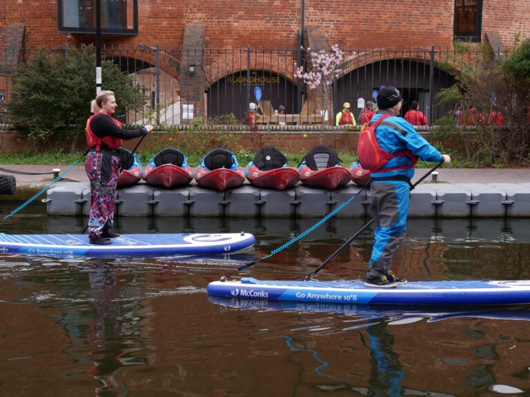A landscape image showing two people stand up paddle boarding on canal water. Behind them is a grey pontoon with a line of red and blue kayaks.