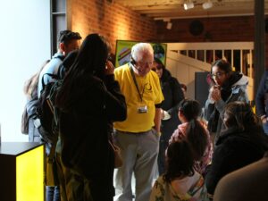 A landscape image showing someone in a yellow shirt and with a headset talking to a group of people and children.