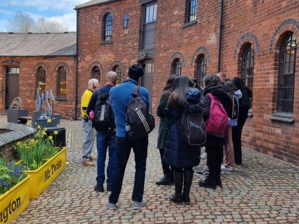 A landscape image showing a group of people outdoors, stood on cobbles listening to a tour. They are stood outdoors.