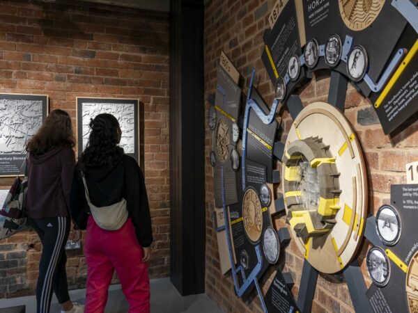 A landscape image showing a large wooden circular display on a red bricked wall. There are two people looking at it.