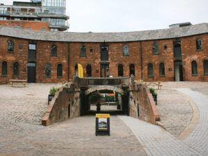 A landscape image looking into a cobbled courtyard. In front, there is a sign and a path behind it leading down under an arched tunnel. Around the side there is a grey path leading up to the red bricked, curved building at the back of the image. In the background, there are high rise buildings.