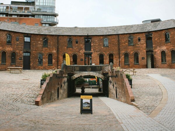 A landscape image looking into a cobbled courtyard. In front, there is a sign and a path behind it leading down under an arched tunnel. Around the side there is a grey path leading up to the red bricked, curved building at the back of the image. In the background, there are high rise buildings.