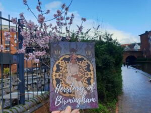 A landscape image showing a hand in the middle holding a piece of paper with the words 'Herstory of Birmingham '. To the left of the paper is cherry blossom, pink against metal grey bars. And to the right of the paper is a green bush, canal towpath and canal water. In the distance is a arched bridge. It is a blue sky day.