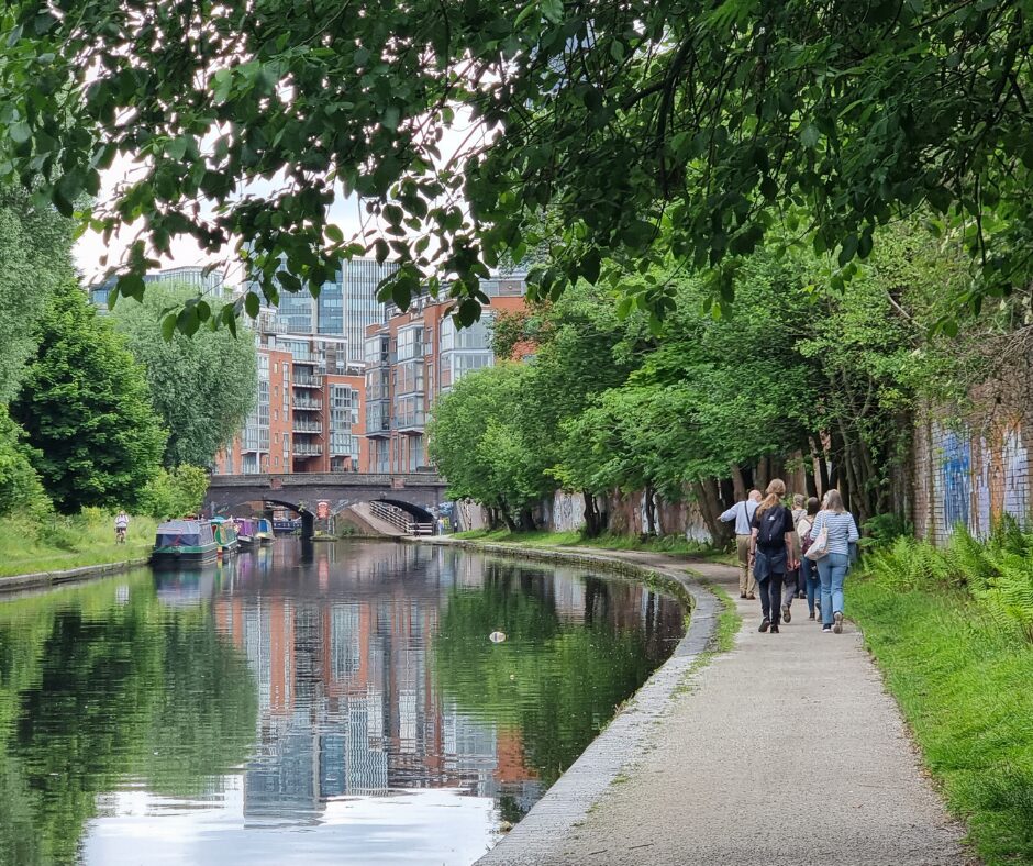 A landscape image showing a canal to the left of the image and to the right of the image is a canal towpath. Walking away from the camera along the path are a group of people. Around the image, along the bank of the canal and towards the group are lots of green shrubbery and plants. In background are some buildings, reflected in the canal.