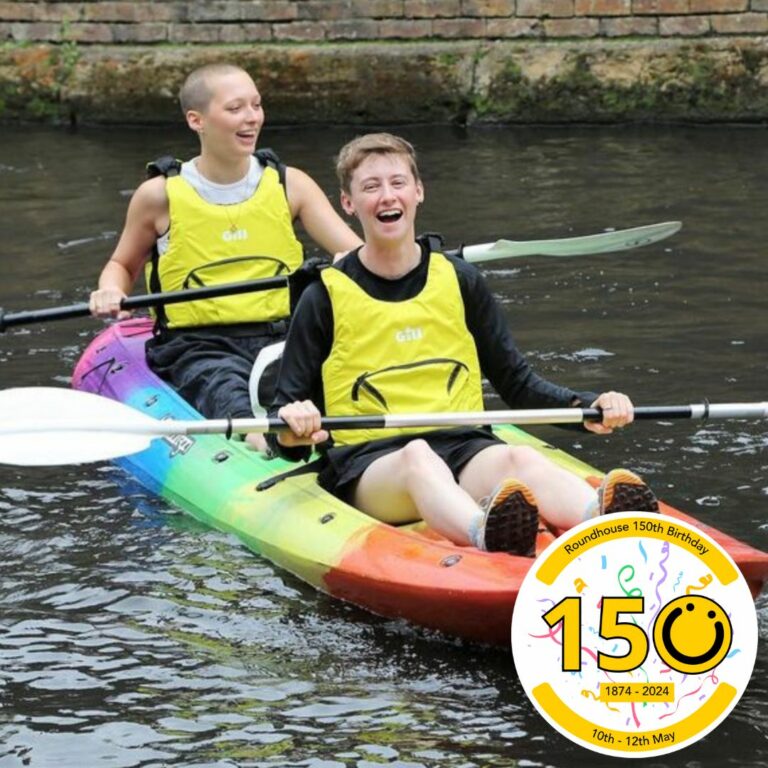 A square image showing two people in one rainbow coloured kayak on canal water. They are both wearing yellow buoyancy aids. In the bottom right of the image is a graphic logo with the number 150, the 0 of the logo is a yellow smiley face logo. The numbers 1 and 5 both have a black outline.