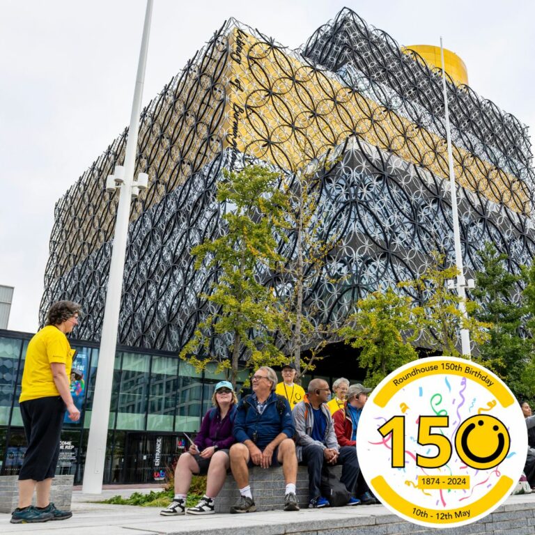 A square image showing a large yellow and grey building. In front of the building are a group of people sat down and in front of them is someone wearing a yellow t-shirt. In the bottom right of the image is a graphic logo with the number 150, the 0 of the logo is a yellow smiley face logo.