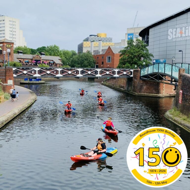 A square image showing six kayakers on canal water. The kayaks are red and blue. To their sides are towpaths. Above them is a black and white metal bridge. In the background and to the right of the image is a grey building with the words, 'SEA LIFE Centre' on and in the background is a yellow and grey building. In the bottom right of the image is a graphic logo with the number 150, the 0 of the logo is a yellow smiley face logo. The numbers 1 and 5 both have a black outline.