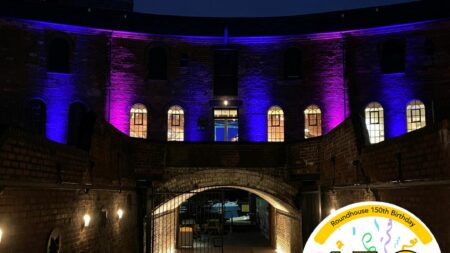 A landscape image showing a large arched tunnel in the centre of the image illuminated with yellow warm light. The flooring is cobbled. Behind and above is a curved red brick walled building. There are six uplighters showing shooting colours of blue and purple. There are multiple arched windows with warm light coming from the inside. In the bottom right of the image is a graphic logo with the number 150, the 0 of the logo is a yellow smiley face logo. The 1, 5 and circular logo are outlined in black.