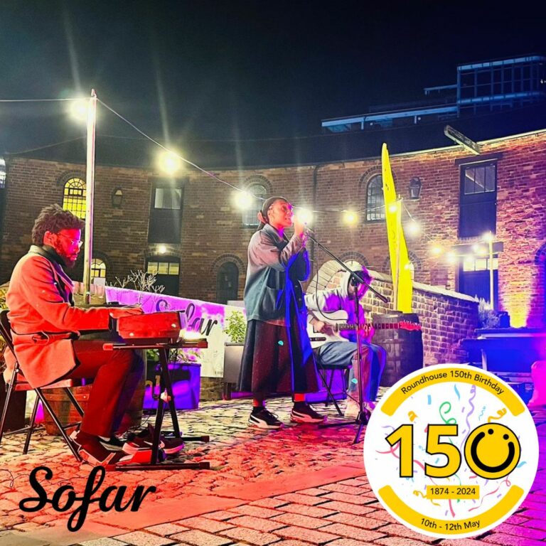 A square image showing someone singing outdoors on cobbled flooring. Behind them are lights and a curved red bricked building. Next to the signer is someone playing a keyboard. In the bottom right of the image is a graphic logo with the number 150, the 0 of the logo is a yellow smiley face logo. To the bottom left of the image is a black font logo saying the name 'Sofar'.