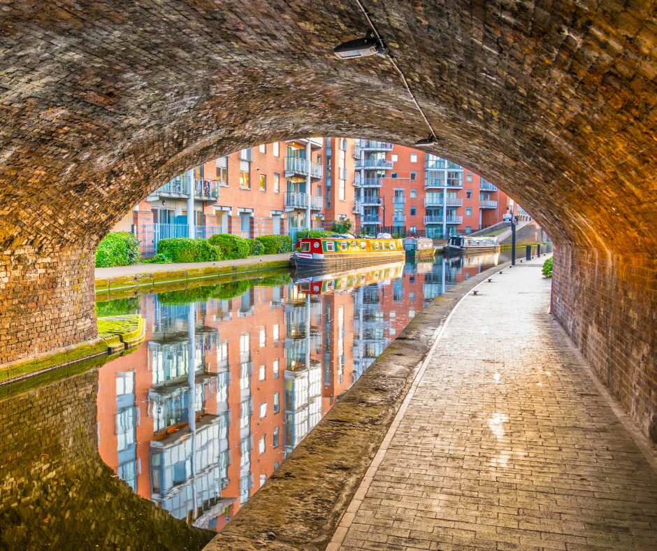 A landscape image showing the view of a canal and path through an arched tunnel. The tunnel is above, and to the left of the image is a canal and to the right is a towpath curving around. On the canal are two canal boats, the front is red, blue and yellow, and the boat behind is darker blue all over. In the background through the tunnel are apartment blocks. The boats and blocks are reflected in the canal water.