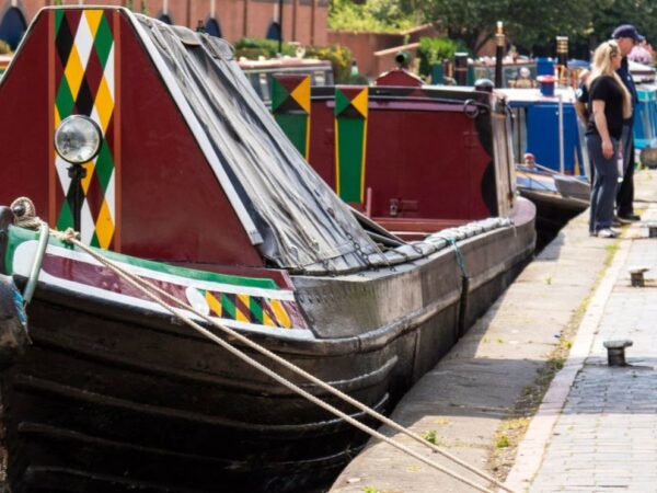 A landscape image showing a burgundy canal boat with black, green, white, burgundy and yellow diamond pattern on the front. The boat is moored up to a path. In the top right of the image are two people stood on the path.