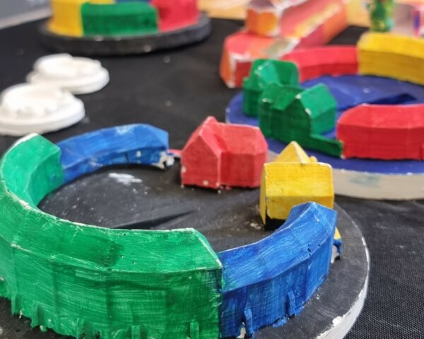 A landscape image showing some small models of a curved building. The models are painted green, blue, red, yellow and black and are on a black table.