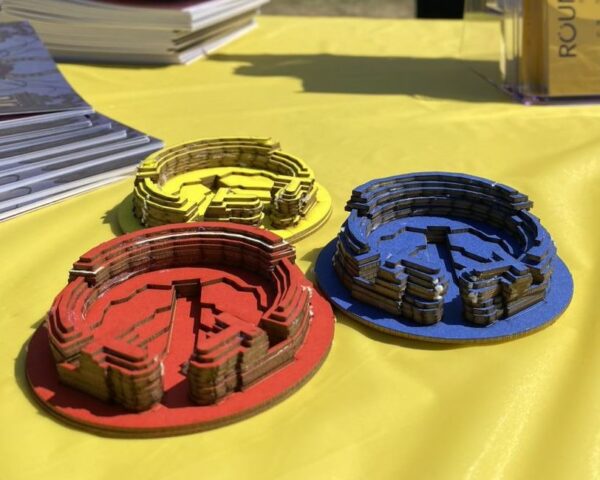 A landscape image showing some small models of a curved building made out of card. One is red, another is blue and another is yellow. The three models are on a yellow table and in the background are a stack of booklets.