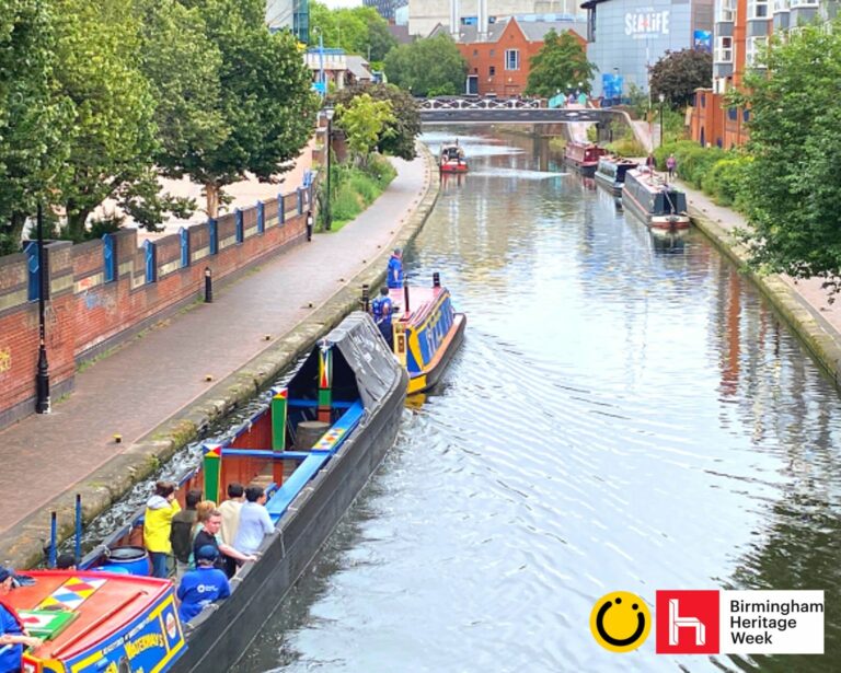 A landscape image showing a view of a canal. Either side of the canal is a path and, on the right, there are people walking along it. On the bottom left part of the image and on the canal is a red roofed and blue and yellow sided canal boat pulling a blue, black, green, red and yellow painted open air boat with 7 people stood on it. In the distance is a black and white metal bridge over the canal and other canal boats on the water. In the bottom right of the image is a yellow smiley face logo and a red square with the letter h in.