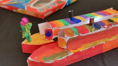 A landscape image showing two small paper canal boat models that have been painted mostly red. In the background is a packet of paints. In the bottom right of the image is a yellow smiley face logo and a red square with the letter h in.