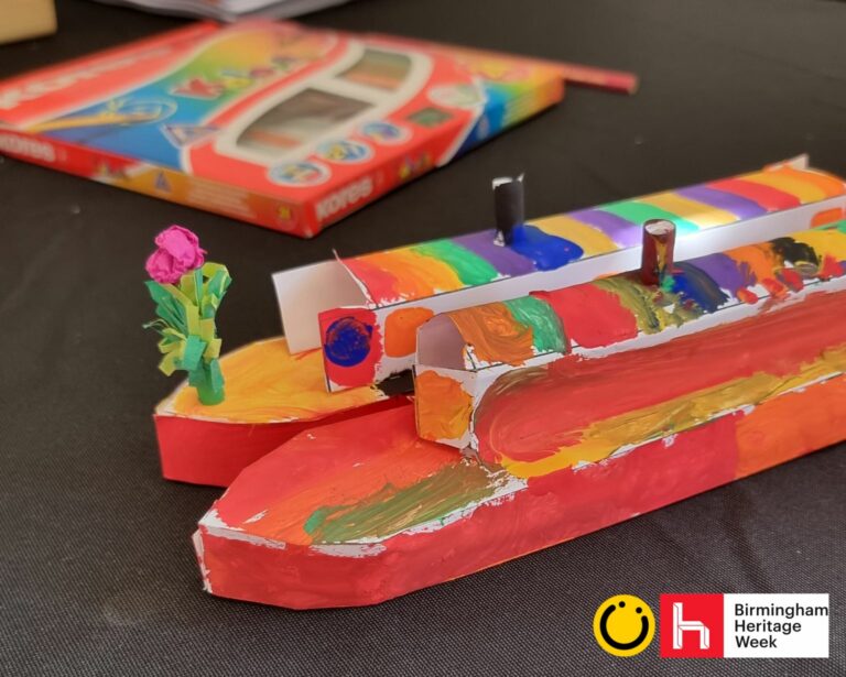 A landscape image showing two small paper canal boat models that have been painted mostly red. In the background is a packet of paints. In the bottom right of the image is a yellow smiley face logo and a red square with the letter h in.